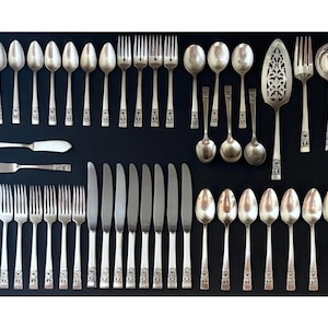 May include: An assortment of vintage silverware, including spoons, forks, knives, and serving utensils, arranged on a black surface. The silverware has ornate detailing on the handles and a polished, reflective finish. A complete set for formal dining.