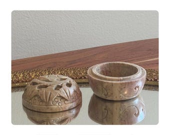 May include: Two beige stone containers. One is a round box with carved floral and leaf designs and cut-out holes. The other is a small, round bowl with a decorative swirl pattern. Both are on a mirrored surface.