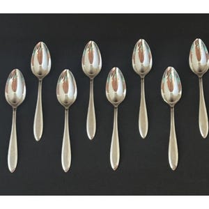 May include: Eight silver spoons arranged on a black surface. The spoons have a classic design with oval bowls and long, slender handles. The spoons are reflective, showing the surrounding environment.