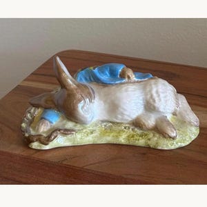May include: A ceramic figurine of a brown rabbit lying down on a textured green base. The rabbit is wearing a blue jacket and is positioned in a resting pose with its head tucked down. The piece features a glossy finish and detailed fur texture.