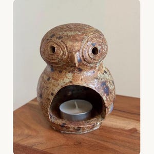 May include: A brown ceramic owl-shaped candle holder with a speckled glaze finish. The owl features large circular eyes and a central hollow opening in the body that contains a small, round white tea light candle with a silver metal casing.
