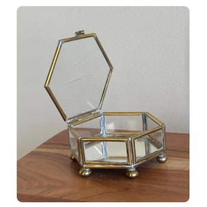 May include: A hexagonal glass jewelry box with a hinged lid, framed in gold-colored metal. The box is open, revealing a mirrored interior and rests on small, rounded feet. The box is displayed on a wooden surface.
