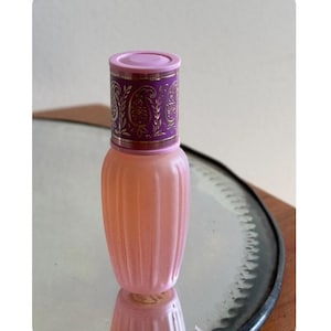 May include: A pink, fluted glass perfume bottle with a decorative purple and gold cap. The bottle is reflected in a mirrored surface, showcasing its symmetrical design. The cap features an ornate paisley pattern.