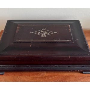 May include: A dark brown wooden box with a rectangular shape and a decorative floral design on the lid. The box has a slightly distressed appearance, suggesting age or wear. The box is sitting on a wooden surface.