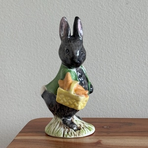 May include: A ceramic figurine of a black rabbit wearing a green jacket, holding a yellow basket filled with orange carrots. The rabbit stands on a base of green grass. The figurine is a decorative Easter or spring-themed item.