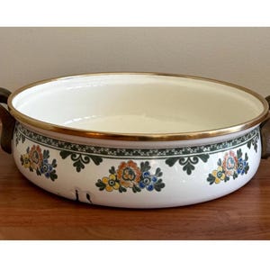 May include: White enamel serving dish with floral pattern and wooden handles. The dish has a gold rim and a decorative band of flowers in shades of blue, yellow, and red. A vintage kitchenware item.