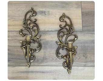 May include: Pair of ornate, antique-style wall sconces in a bronze finish. Each sconce features a floral design and scrolling details, with a candle holder. The sconces are mounted on a wooden surface.