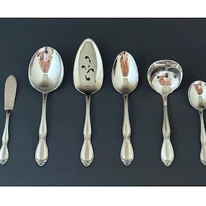 Puede incluir: Un juego de seis utensilios de servir de color plateado, que incluye un cuchillo de mantequilla, dos cucharas, una cuchara ranurada, un cucharón y una cuchara más pequeña. Los utensilios están dispuestos sobre una superficie gris oscuro.