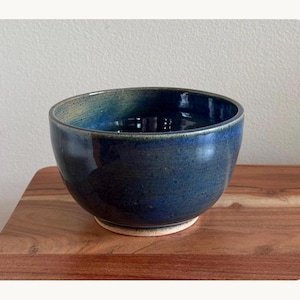 May include: A round ceramic bowl with a deep blue glaze that transitions to lighter teal and green tones near the rim. The bowl features a smooth, glossy finish with subtle dark speckles throughout the surface and a plain, unglazed light beige base.