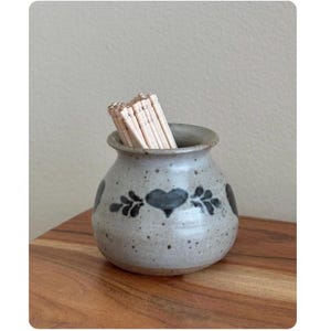 May include: A small, speckled ceramic jar with a heart and leaf design, filled with wooden toothpicks. The jar is gray with dark blue accents and sits on a wooden surface.