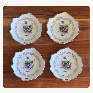 May include: Four white porcelain dishes shaped like leaves with scalloped edges. Each dish features a central cluster of purple and white violet flowers with green leaves, surrounded by a delicate border of small grey foliage patterns.