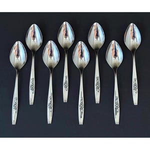 May include: A set of eight silver spoons arranged on a black surface. The spoons have a classic design with a floral pattern on the handles. The spoons are reflective, showing the surrounding environment.