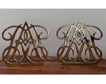 May include: A pair of gold-colored metal bookends featuring an intricate, symmetrical openwork design that resembles a stylized letter A. The metallic surfaces have a polished, reflective finish, and the structures are shaped with curved lines.