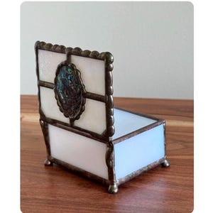 May include: A small, decorative stained glass box with a hinged lid. The box features a white and iridescent abalone shell design, framed by a dark metal border. The box is approximately 3 inches long and 2 inches wide.