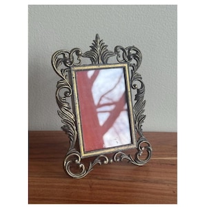 May include: A gold-toned metal picture frame with an ornate design. The frame is standing on a wooden surface. The frame is empty, but there is a faint shadow of a tree branch inside.