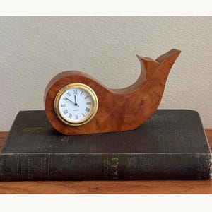 May include: A whale-shaped wooden desk clock featuring a polished brown wood grain finish. The clock face is circular with a gold-toned metal rim, a white background, black Roman numerals, and black hour, minute, and second hands.