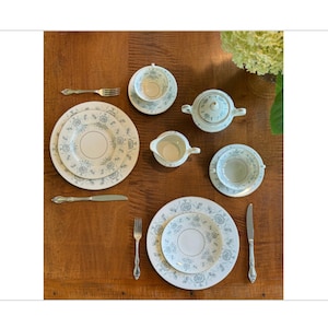 May include: A set of white porcelain teacups and saucers with a delicate floral pattern in shades of blue and green. The set includes a creamer, sugar bowl, and two teacups and saucers. The teacups and saucers are arranged on a wooden table with silver cutlery.