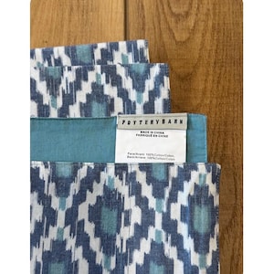 May include: Close-up of folded blue and white patterned fabric with a teal border. The fabric has a geometric design. A tag reads "POTTERY BARN MADE IN CHINA FABRIQUE EN CHINE". The fabric is made of 100% cotton.