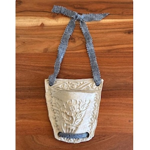 May include: A cream-colored, textured ceramic planter with a floral design, suspended by a blue ribbon. The planter has a pocket shape and is designed to hang. The ribbon is a textured, woven material.