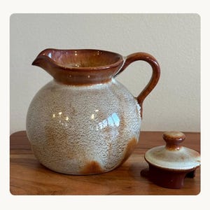 May include: A round ceramic pitcher with a glossy brown rim and handle, featuring a textured, speckled cream and beige body. The pitcher has a wide spout and a matching removable lid with a small knob handle, showcasing a consistent glaze.