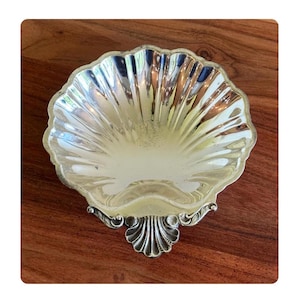 May include: A silver-toned metal dish shaped like a scallop shell with a polished, reflective surface and deep, radiating fluted ridges. The base features a decorative, smaller shell-shaped foot with ornate scrollwork details at the bottom edge.
