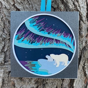 May include: A paper craft ornament featuring a circular design of the aurora borealis in shades of blue, purple, and green with two white polar bears standing on a snowy landscape. The ornament is hanging from a blue ribbon.