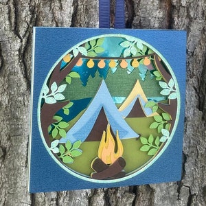 May include: A blue papercraft ornament depicting a camping scene with two tents, a campfire, and string lights hanging from trees.