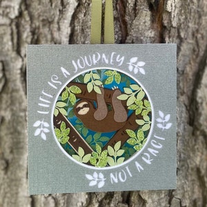 May include: Square wall art with a sloth illustration hanging from a tree branch, surrounded by green leaves and the text "LIFE IS A JOURNEY NOT A RACE". The art is on a textured gray background and has a green ribbon for hanging.