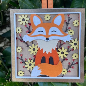 May include: A square, layered paper art piece featuring a fox. The fox is orange and white, surrounded by yellow flowers and red berries. The artwork is framed in gold and has an orange ribbon for hanging.