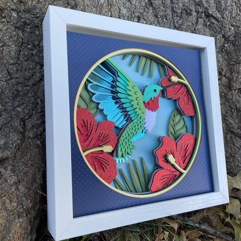 May include: A framed, layered paper art piece featuring a hummingbird with turquoise wings, a red throat, and green body, hovering near red hibiscus flowers. The artwork is set against a blue background with a gold border, all within a white frame.