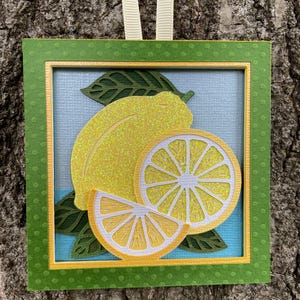 May include: A square wall hanging featuring a layered paper craft of a whole lemon, a lemon slice, and a lemon wedge with glittery yellow surfaces. The design includes dark green leaves on a light blue background, framed by a green polka dot border.