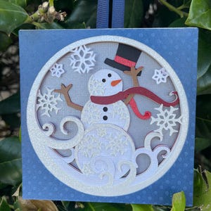 May include: A square, blue Christmas ornament with a snowman design. The snowman has a black top hat, a red scarf, and brown arms. White snowflakes and a glittery border surround the snowman. A blue ribbon is attached for hanging.
