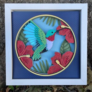 May include: A framed, layered paper art piece featuring a hummingbird and hibiscus flowers. The hummingbird has blue wings, a green body, and a red throat. The red hibiscus flowers have glitter accents, set against a blue background.