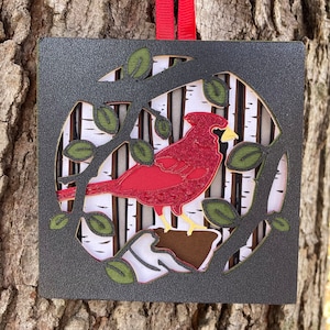 May include: A square, layered ornament featuring a red cardinal bird perched on a branch. The bird is surrounded by green leaves and a birch tree background, all within a dark gray frame. A red ribbon is attached for hanging.