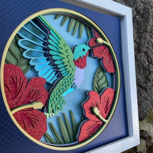 May include: A framed, layered paper art piece featuring a hummingbird and hibiscus flowers. The hummingbird has green, blue, and red details. The flowers are red with glitter accents, set against a blue background. The artwork is in a white frame.