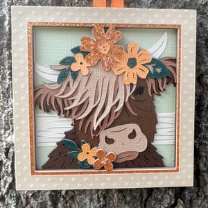 May include: A layered paper art piece featuring a Highland cow with a floral crown. The cow is brown with long, shaggy fur and a light-colored muzzle. The frame is off-white with a dotted pattern and a copper-colored inner border. An orange ribbon is attached for hanging.
