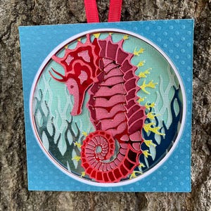 May include: A layered paper ornament featuring a pink seahorse design. The seahorse is set against a light blue background with stylized seaweed and a white circular frame. The ornament has a light blue square border with a dotted pattern and a red ribbon for hanging.