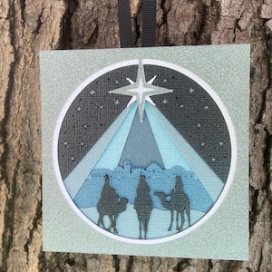 May include: A silver glitter card with a blue and white paper cut-out design of the three wise men following a star. The star is a white paper cut-out with a blue center.