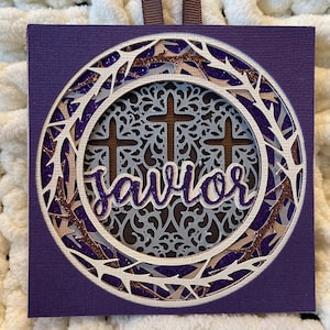 May include: A purple card with a white circle containing three crosses and the word "Savior" in purple glitter. The circle is surrounded by a white crown of thorns.