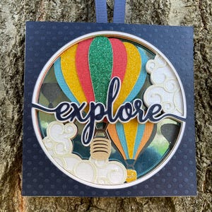 May include: A square, navy blue card with white polka dots features a round, layered, paper craft design of hot air balloons with the word "explore" in a black, cursive font.
