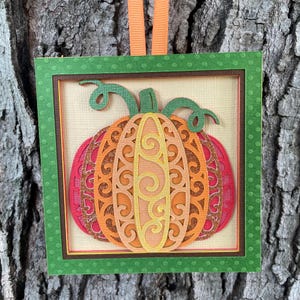 May include: A layered paper pumpkin ornament with a green frame and orange ribbon. The pumpkin features intricate cut-out designs in shades of orange, red, and yellow. The background is a light beige color.