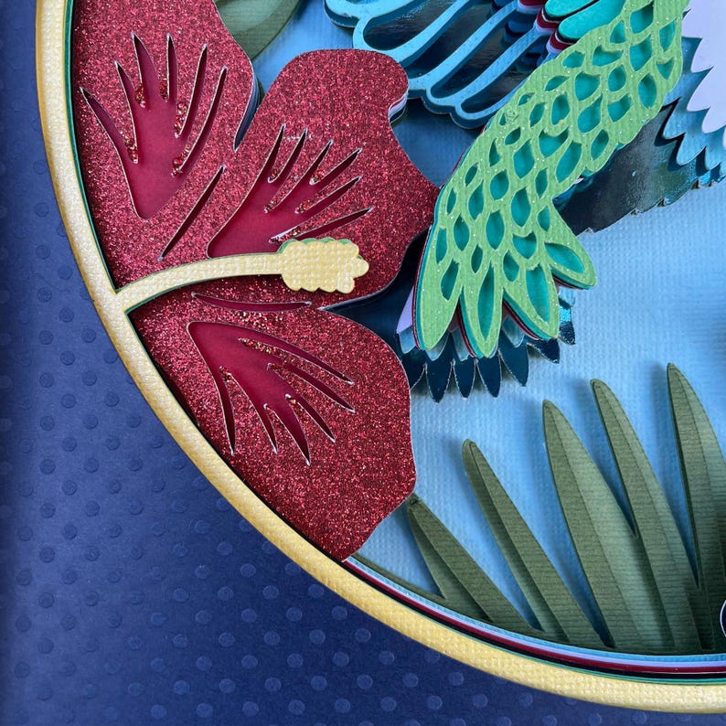 May include: A layered paper art piece featuring a red glitter hibiscus flower with yellow accents and green leaves. A green and blue bird is also visible. The artwork is set against a blue background.