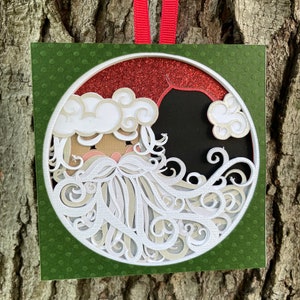 May include: A layered paper Christmas ornament featuring Santa Claus. The design includes a red glitter hat, white beard, and a green background with a polka dot pattern. A red ribbon is attached for hanging.