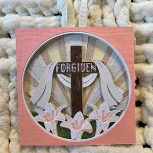 May include: A pink square card with a circle cut out in the center. The circle contains a brown wooden cross with the word "FORGIVEN" written on it. The cross is surrounded by white paper flowers and a white paper background with a sunburst design.