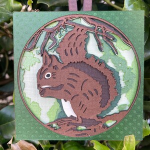 May include: A square, green ornament with a detailed, layered design of a squirrel in a forest setting. The squirrel is brown with white accents, set within a circular frame. A brown ribbon is attached for hanging.