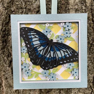 May include: A square hanging ornament featuring a layered paper butterfly with black wings and blue glitter accents. The butterfly is set against a yellow and white lattice background, surrounded by small light blue flowers and green leaves.