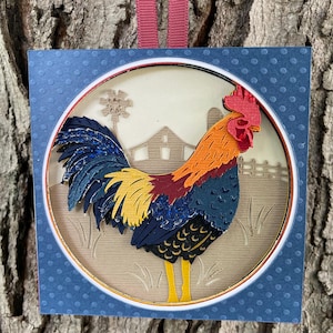 May include: A layered paper art piece featuring a colorful rooster with a red comb, orange and blue feathers, and yellow legs. The rooster is set against a farm scene with a barn, windmill, and fence, framed by a blue dotted border.