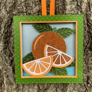 May include: A square layered paper ornament featuring a glittery orange fruit and two sliced orange segments. The design includes dark green die-cut leaves against a light blue background, framed by a green polka-dot border with an orange ribbon.