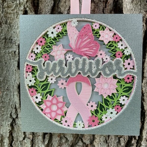 May include: A layered paper craft ornament with a gray background, featuring a pink ribbon, butterfly, and flowers. The word "Survivor" is displayed in silver glitter. A pink ribbon hangs from the top.