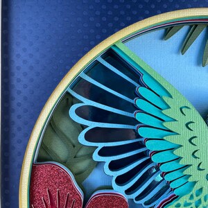 May include: Close-up of a layered paper art piece featuring a stylized bird or insect wing. The artwork uses shades of blue, teal, green, and red with glitter accents. The background is a dark blue with a dotted pattern.